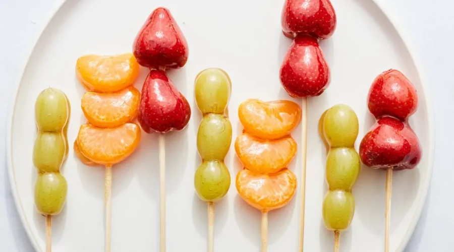 Espetinhos de frutas coloridas cobertos com uma camada brilhante de caramelo, dispostos em um prato branco.