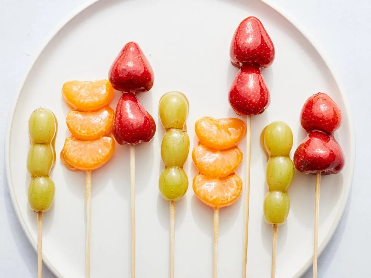 Espetinhos de frutas coloridas cobertos com uma camada brilhante de caramelo, dispostos em um prato branco.