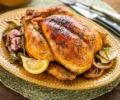 Frango assado inteiro com pele dourada, servido sobre uma cama de cebolas caramelizadas, alho assado e limão siciliano em gomos, em uma assadeira rústica.