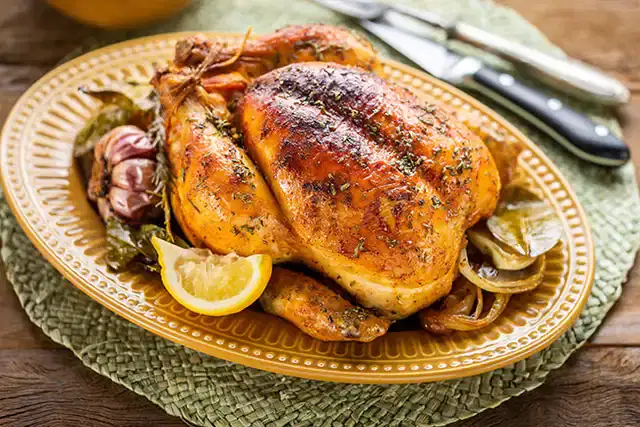 Frango assado inteiro com pele dourada, servido sobre uma cama de cebolas caramelizadas, alho assado e limão siciliano em gomos, em uma assadeira rústica.
